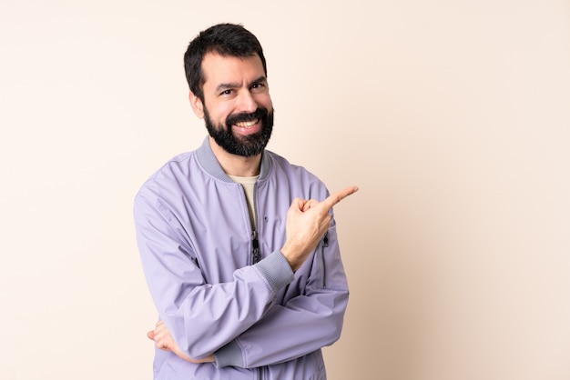 Homme de race blanche avec barbe portant une veste sur le mur pointant vers le côté pour présenter un produit