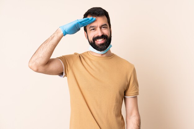 Homme de race blanche avec barbe protégeant avec un masque et des gants sur le mur saluant avec la main avec une expression heureuse