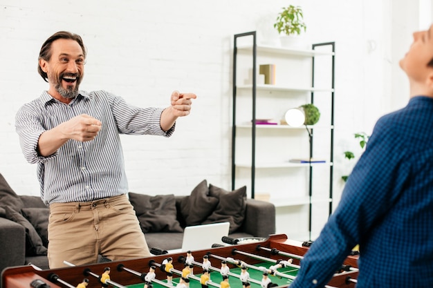 Homme de race blanche barbu excité en montrant son fils adolescent et en riant après avoir joué au baby-foot avec lui