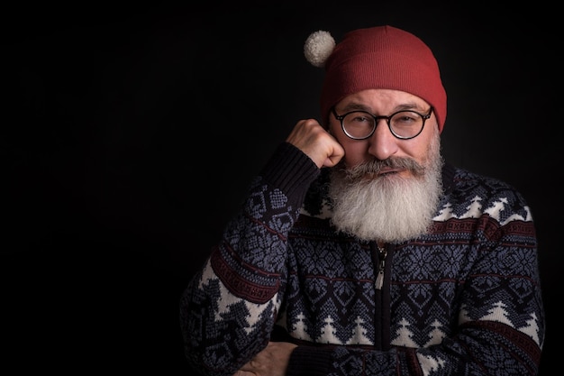 Homme de race blanche barbu mature regarde sournoisement à huis clos avec des lunettes sur son visage homme en chapeau de père noël rouge fond noir