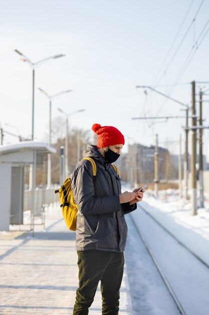 homme de race blanche dans une veste sombre et un chapeau rouge dans une gare en attente d'un train et de textos