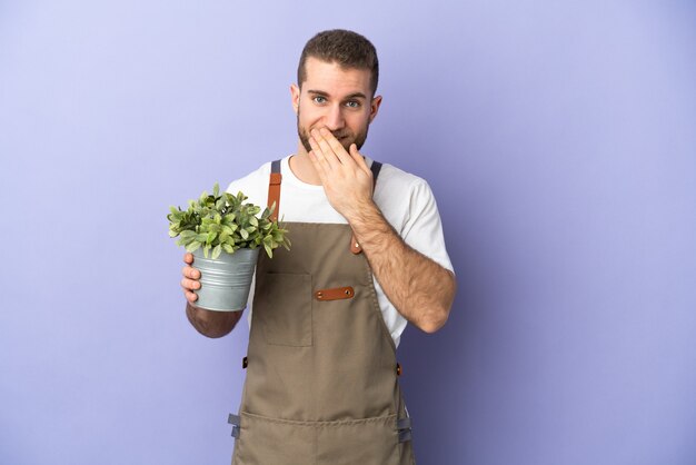 Homme de race blanche jardinier tenant une plante isolée sur mur jaune heureux et souriant couvrant la bouche avec la main