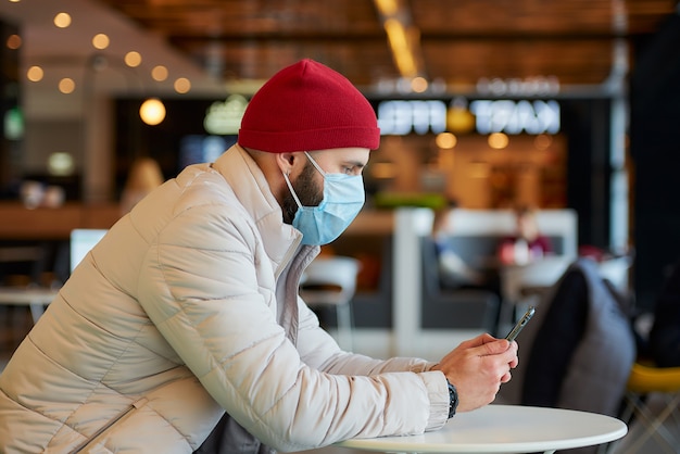 Un homme de race blanche avec un masque chirurgical sur le visage à l'aide d'un smartphone dans le centre commercial.