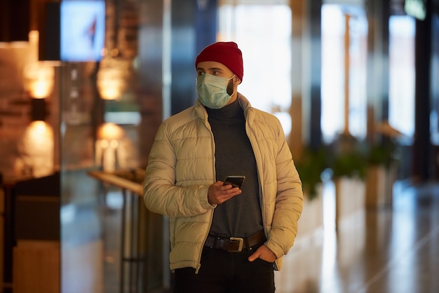 Un homme de race blanche avec un masque chirurgical sur le visage à l'aide d'un smartphone dans le centre commercial.