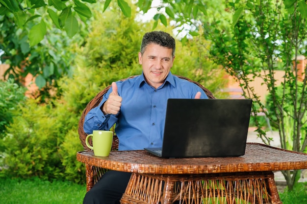 Homme de race blanche mature souriant buvant du café et montrant les pouces vers le haut avec les doigts à un partenaire commercial via un ordinateur portable dans le jardin de la maison. Concept d'entreprise à distance