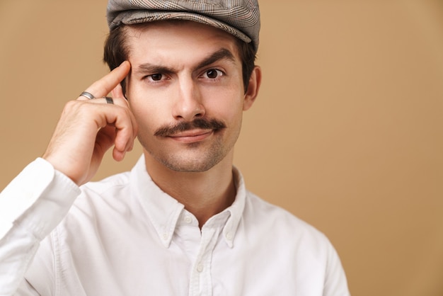 homme de race blanche à moustache portant un chapeau regardant à l'avant et touchant sa tempe isolée sur beige