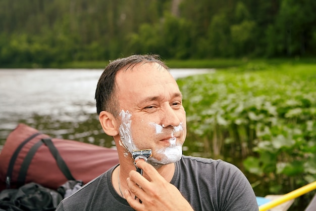 Un homme de race blanche se rase le visage à l'aide de mousse et de lame de rasoir le matin près de la rivière pendant ses vacances.