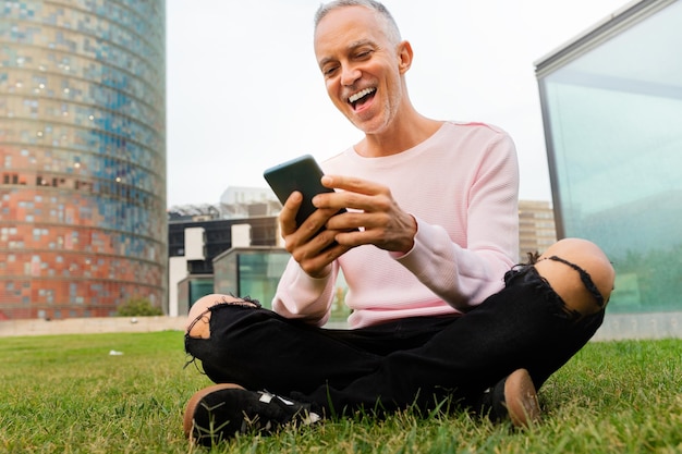 Homme de race blanche souriant assis sur l'herbe dans le parc public de la ville en regardant le téléphone portable. Concept de médias sociaux et de technologie.