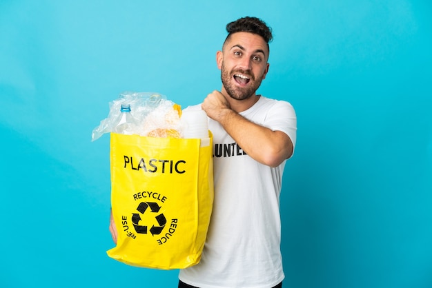 Homme de race blanche tenant un sac plein de bouteilles en plastique à recycler isolé sur fond bleu célébrant une victoire