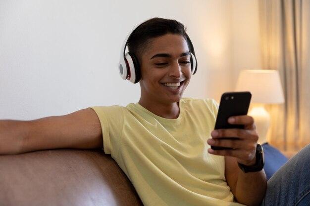 Homme de race mixte assis sur un canapé portant des écouteurs souriant et utilisant un smartphone