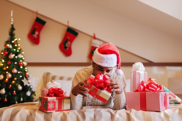 Homme de race mixte avec chapeau de père Noël sur la tête allongée sur le lit et essayant d'ouvrir le cadeau de Noël.