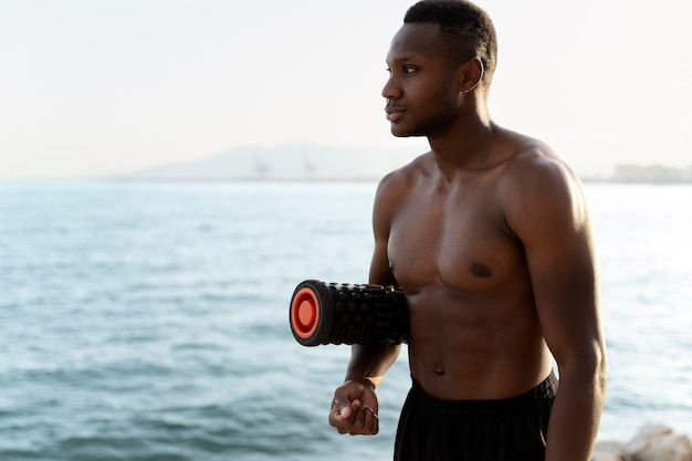 Homme de race mixte avec torse nu debout au bord de la mer avec un tapis de yoga et en détournant les yeux