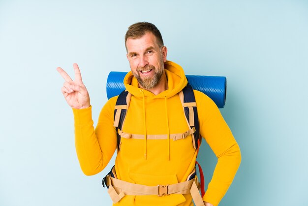 Homme de randonneur senior isolé sur mur bleu joyeux et insouciant montrant un symbole de paix avec les doigts.