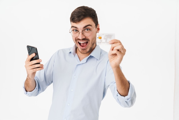 homme ravi à lunettes tenant un téléphone portable et une carte de crédit isolé sur un mur blanc