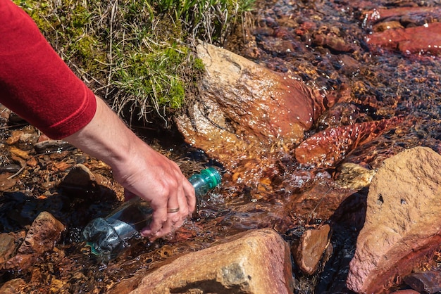 Un homme recharge l'eau d'un ruisseau de montagne en plein soleil Un touriste remplit une bouteille d'eau minérale en journée ensoleillée La main avec la bouteille dans un ruisseau d'eau claire en plein soleil Recharge d'eau pendant la randonnée