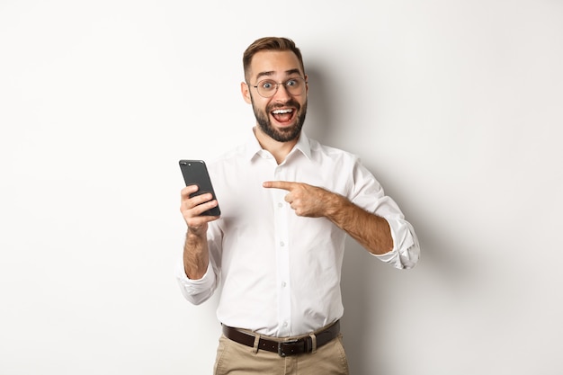 Homme à la recherche de doigt excité et pointé au téléphone mobile, montrant une bonne offre en ligne, debout sur fond blanc.