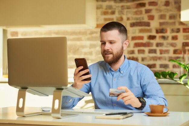 Un homme recherche des ventes en ligne sur un smartphone, tenant une carte de crédit à la main devant l'ordinateur à la maison