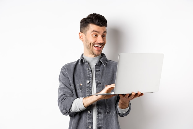 Homme regardant une vidéo drôle sur ordinateur portable, riant à l'écran d'ordinateur et souriant joyeux, debout sur fond blanc.
