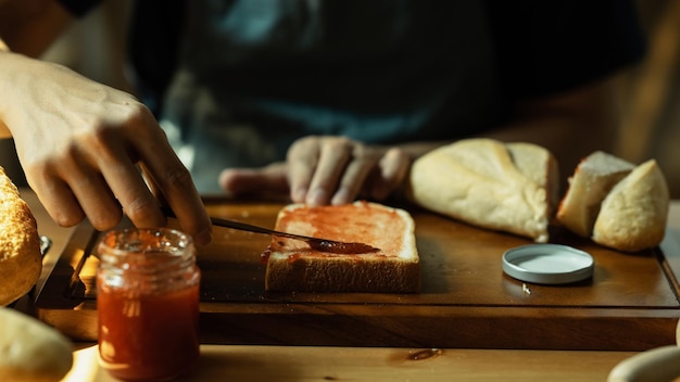 Homme répandant de la confiture de framboises sucrées sur du pain pendant le petit-déjeuner ou le brunch dans la cuisine