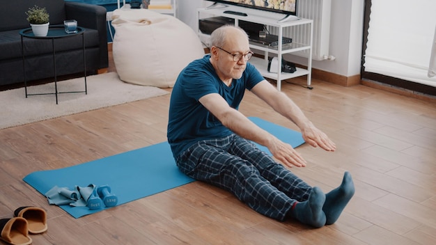 Homme à la retraite faisant de l'exercice physique pour pratiquer la gymnastique et l'activité physique. Personne âgée étirant les bras et les jambes pour un entraînement sain, assise sur un tapis de yoga. Retraité actif