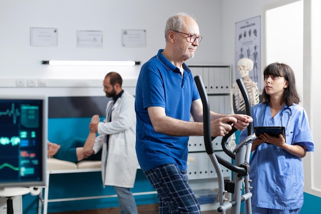 Homme à la retraite faisant de l'exercice physique sur un vélo stationnaire pour récupérer pendant qu'une infirmière tient une tablette. Patient avec entraînement contre les douleurs aux jambes pour le soulagement musculaire et les soins de santé en physiothérapie.