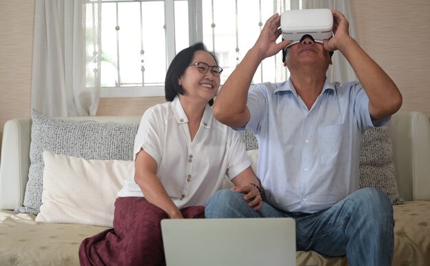 Un homme à la retraite portant un casque VR et sa femme assis côte à côte en train de passer un appel vidéo sur le canapé de la maison.