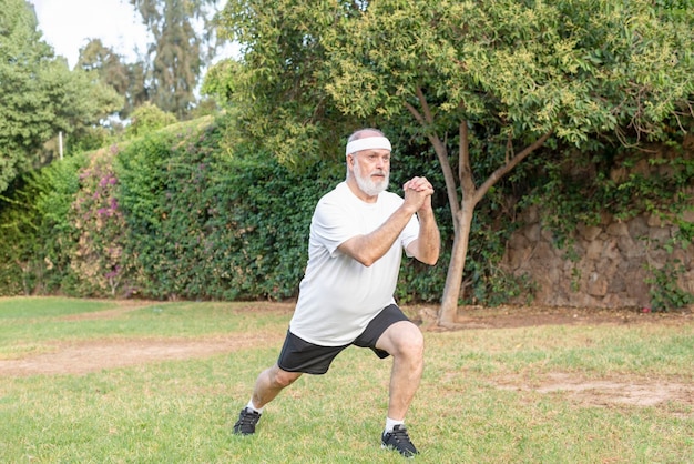 Homme à la retraite en vêtements de sport faisant du squat avant sur une jambe vers l'avant avec les mains jointes dans le parc public Concept de mode de vie actif