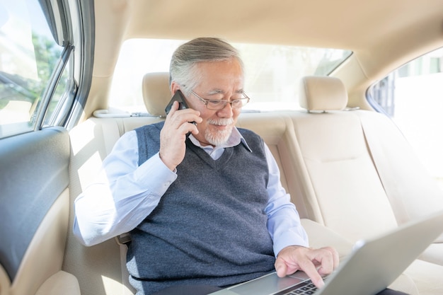 Homme riche senior d'affaires Joueur de commerçant en costume travaillant avec un ordinateur portable et utilisant un téléphone intelligent dans sa voiture, concept de réussite commerciale senior