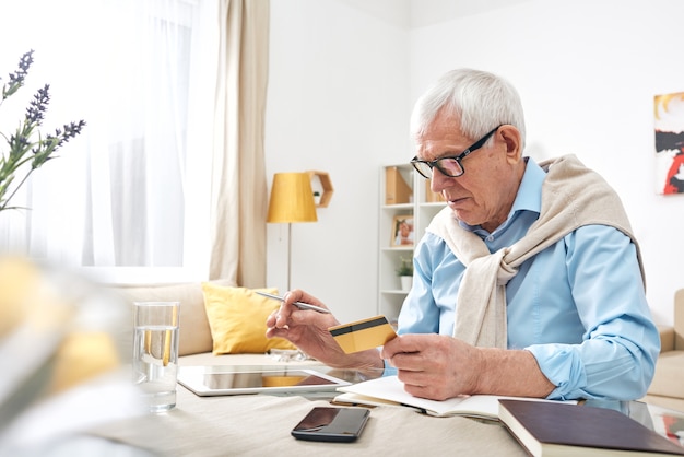 Homme ridé âgé occupé à lunettes à l'aide de tablette numérique tout en vérifiant le solde de la carte de débit à la maison