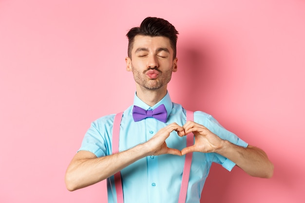 Homme romantique en noeud papillon drôle attendant le baiser, fermez les yeux et les lèvres plissées s'embrassant, montrant le geste du cœur, étant amoureux le jour de la Saint-Valentin, debout sur fond rose.