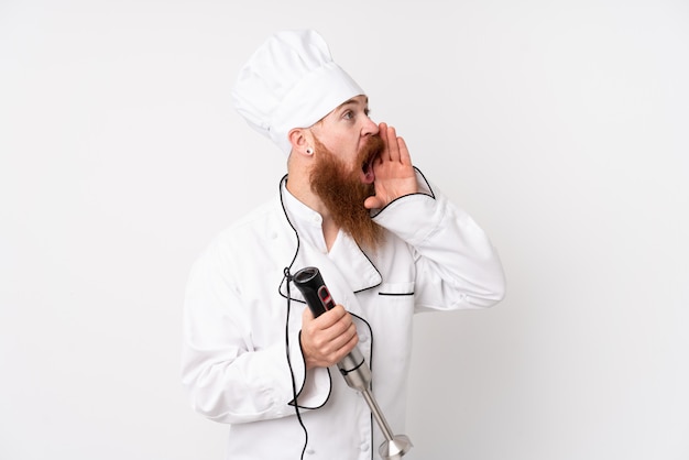 Homme rousse à l'aide d'un mélangeur à main sur un mur blanc isolé criant avec la bouche grande ouverte