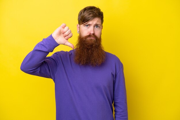 Homme rousse avec barbe isolé sur fond jaune montrant le pouce vers le bas avec une expression négative