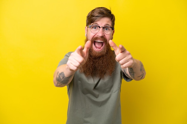 Homme rousse avec barbe isolé sur fond jaune surpris et pointant vers l'avant