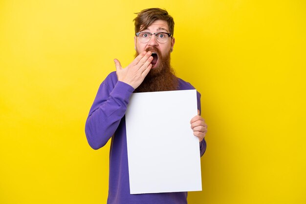 Homme rousse avec barbe isolé sur fond jaune tenant une pancarte vide avec une expression surprise