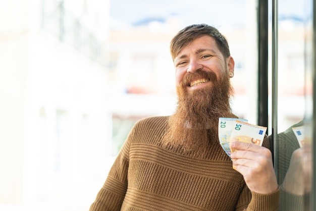 Homme rousse avec barbe prenant beaucoup d'argent à l'extérieur souriant beaucoup