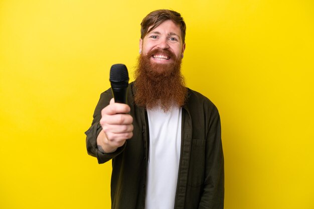 Homme rousse avec barbe ramasser un microphone isolé sur fond jaune avec une expression heureuse