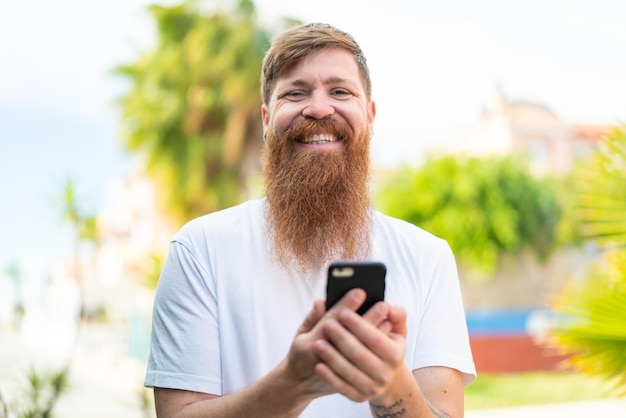 Homme rousse avec barbe regardant la caméra et souriant tout en utilisant le mobile