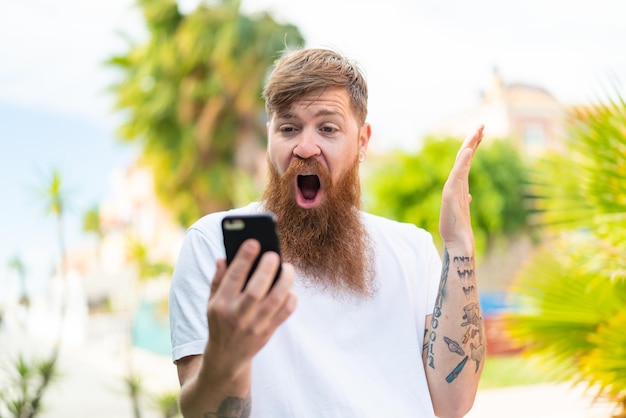 Homme rousse avec barbe regardant la caméra tout en utilisant le mobile avec une expression surprise