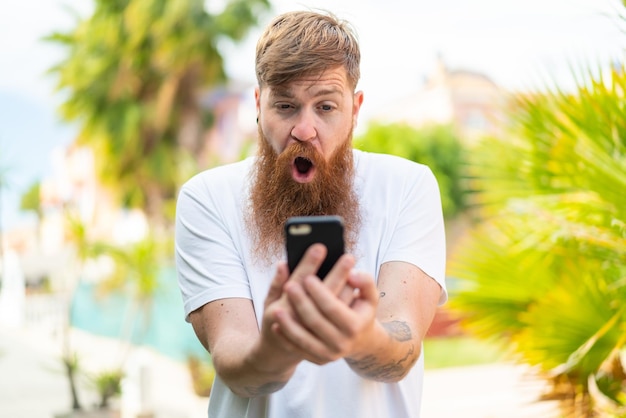 Homme rousse avec barbe regardant la caméra tout en utilisant le mobile avec une expression surprise