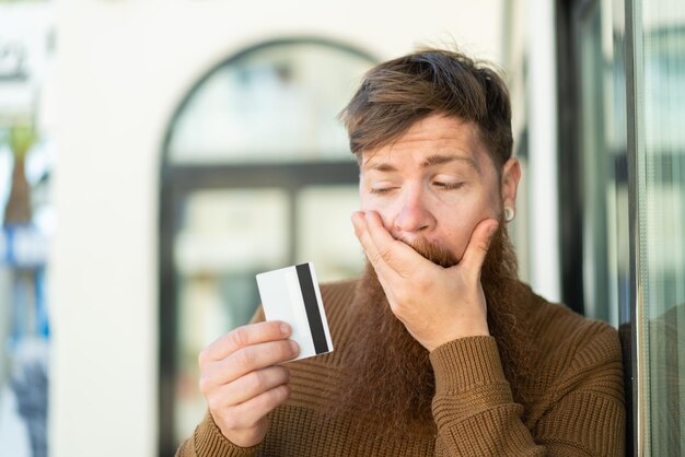 Homme rousse avec barbe tenant une carte de crédit avec une expression surprise