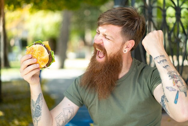 Homme rousse avec barbe tenant un hamburger à l'extérieur célébrant une victoire