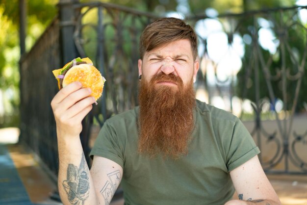 Homme rousse avec barbe tenant un hamburger à l'extérieur avec une expression triste