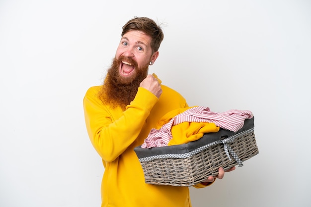 Homme rousse avec barbe tenant un panier à linge isolé sur fond blanc célébrant une victoire