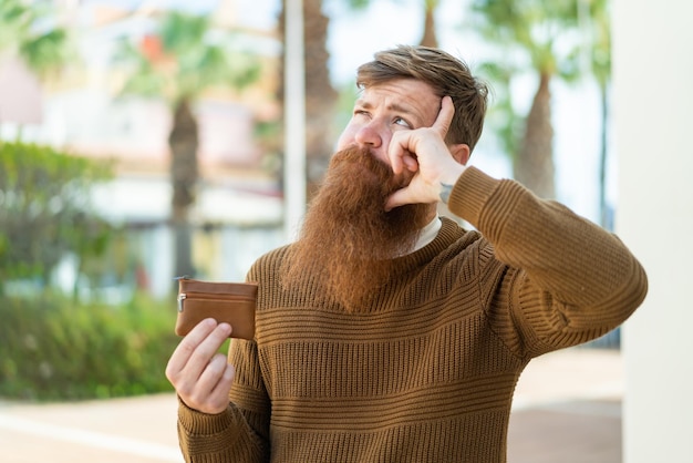 Homme rousse avec barbe tenant un portefeuille à l'extérieur ayant des doutes et avec une expression de visage confuse