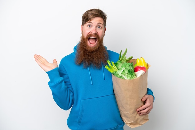 Homme rousse avec barbe tenant un sac d'épicerie isolé sur fond blanc avec une expression faciale choquée