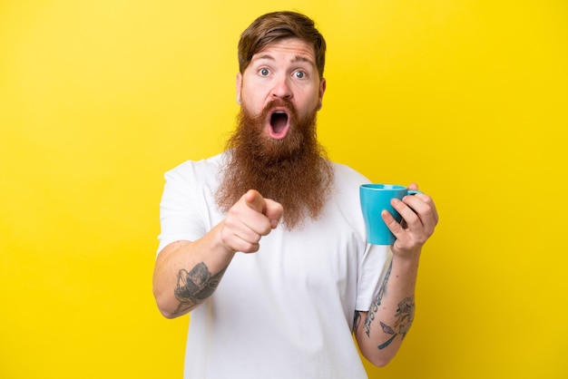 Homme rousse avec barbe tenant une tasse isolée sur fond jaune surpris et pointant vers l'avant