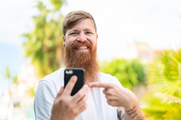 Homme rousse avec barbe utilisant un téléphone portable à l'extérieur et le pointant