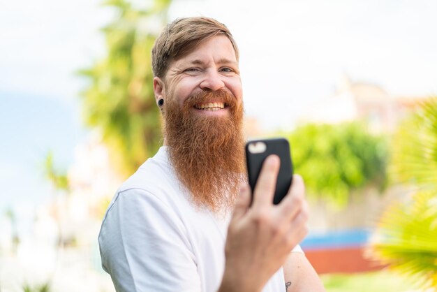 Homme rousse avec barbe utilisant un téléphone portable à l'extérieur souriant beaucoup