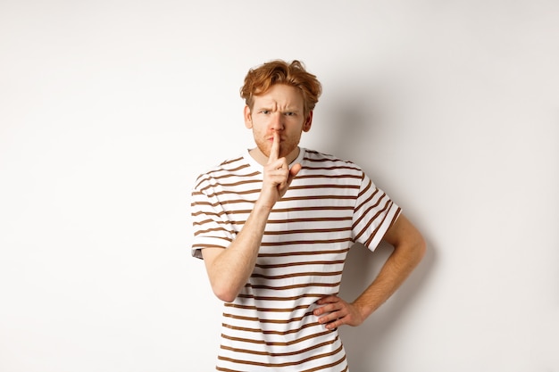 Homme rousse en colère à la recherche de mécontentement et de silence à la caméra, faisant un geste chut, debout sur fond blanc.