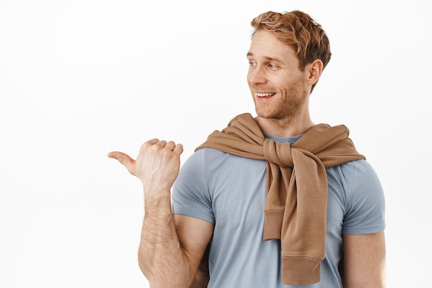 Homme rousse fort et masculin élégant pointant vers la gauche avec un sourire heureux regardant quelque chose de bien avoir une opinion positive sur la promotion du produit debout sur fond blanc